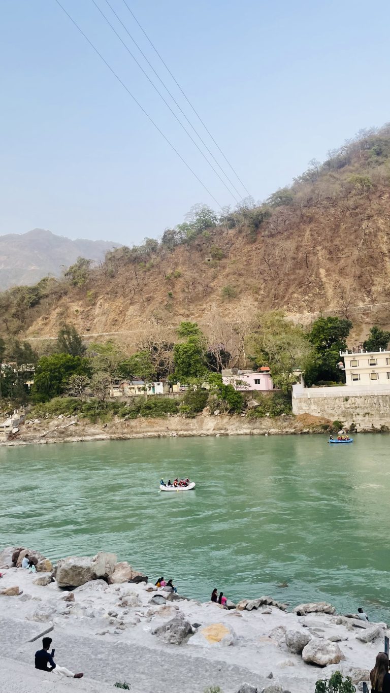 A scenic view of a river with a group of people rafting, surrounded by rocky hills and lush greenery. In the foreground, people sit and walk along the riverbank.