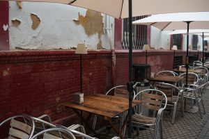 Outdoor café seating area with metal chairs and wooden tables under large umbrellas. The area is set against a red brick wall with peeling white paint.