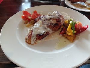 Grilled chicken breast with creamy sauce on a white plate, served with sliced red and yellow bell peppers, tomatoes, and raisins.