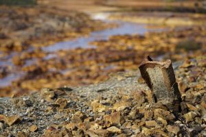 A rusty, broken metal piece of railroad track protrudes from rocky ground in focus, with a blurred background of a stream and rocky landscape.