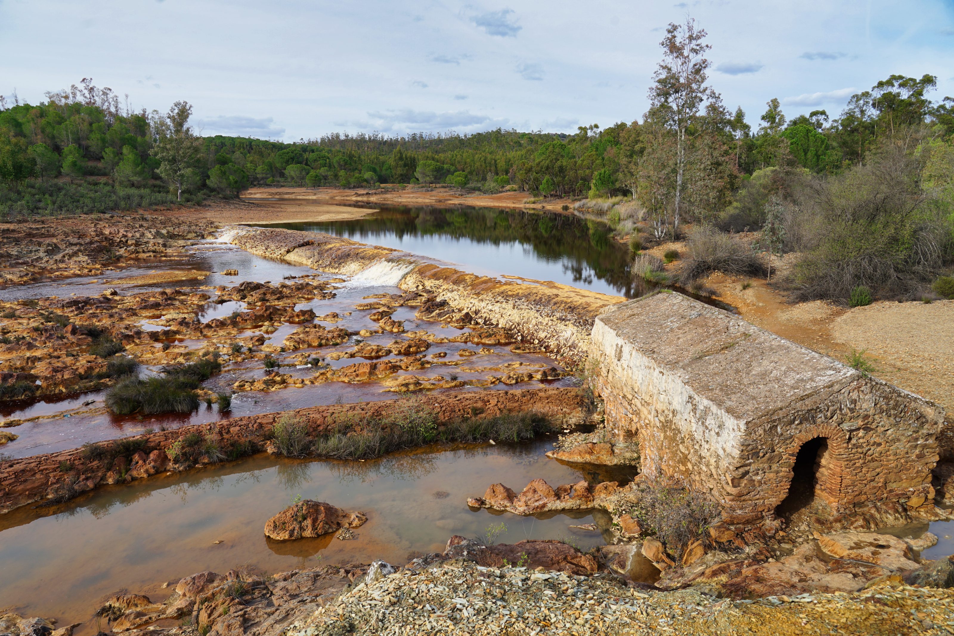 Tinto river flows through a rocky landscape with reddish-brown stones and patches of vegetation. In the foreground, an old stone mill with an arch is partially submerged in water. 