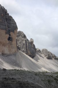 A scenic view of rugged, rocky mountains with steep slopes under a cloudy sky. The terrain is a mix of exposed rock and sparse vegetation. Italian Dolomites