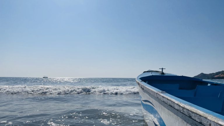 A close-up of a small boat by the shore, with gentle waves and a distant boat on the horizon under a clear blue sky.