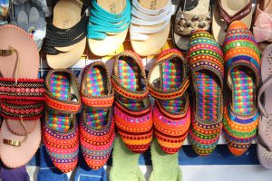 
A display of traditional Indian colorful footwear including vibrant, patterned slip-on shoes and various styles of sandals, arranged on a shelf.