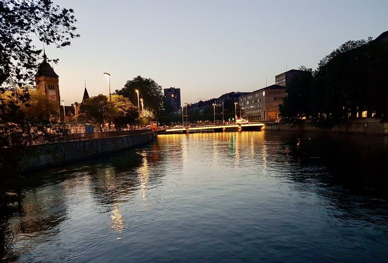 Serene riverside view of Zurich, where the calm waters reflect the beauty of the city’s historic architecture and natural surroundings.