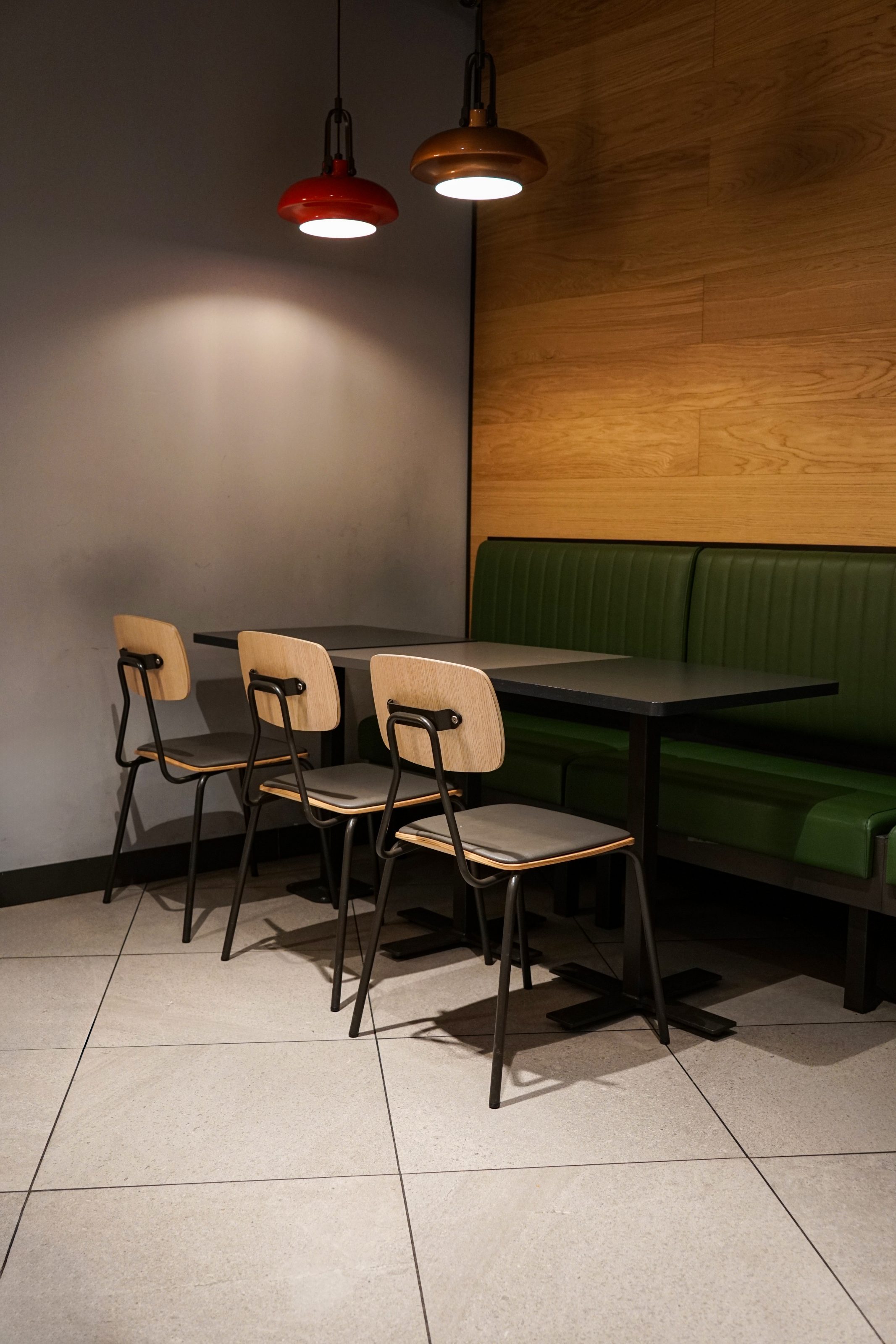 A cozy dining area with a green booth and a black table, accompanied by three wooden chairs. The wall has a wooden panel finish, and two pendant lights, hang above the table.