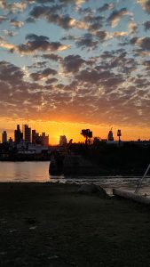 A vibrant sunset illuminates the sky with hues of orange and yellow, casting a silhouette over a city skyline near a body of water. Scattered clouds are tinged with the fading sunlight, and there is a grassy area in the foreground.