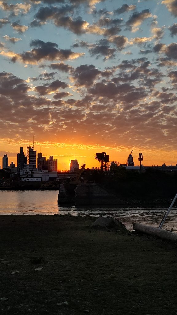 A vibrant sunset illuminates the sky with hues of orange and yellow, casting a silhouette over a city skyline near a body of water. Scattered clouds are tinged with the fading sunlight, and there is a grassy area in the foreground.