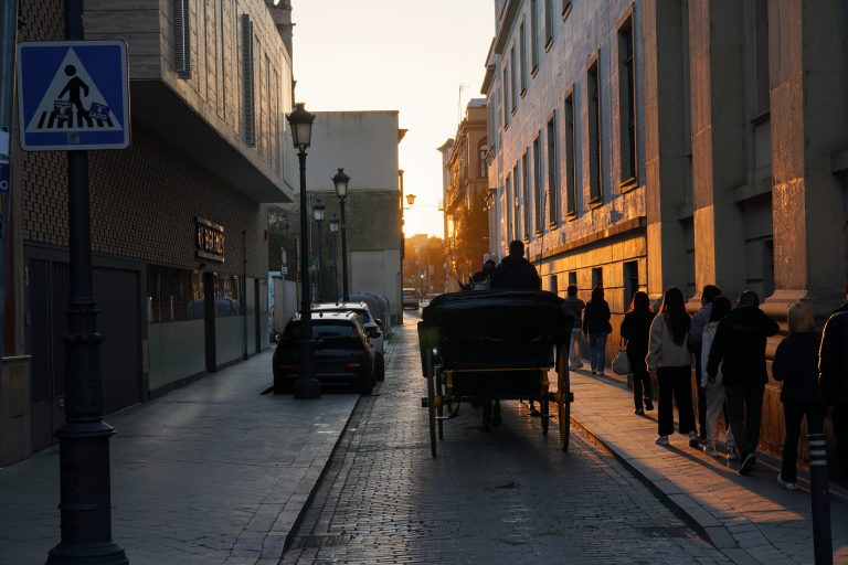 A narrow street at sunset with people walking along the sidewalk on the right, where the buildings are illuminated by the low sun. A horse-drawn carriage travels down the cobblestone road, and parked cars line the left side.