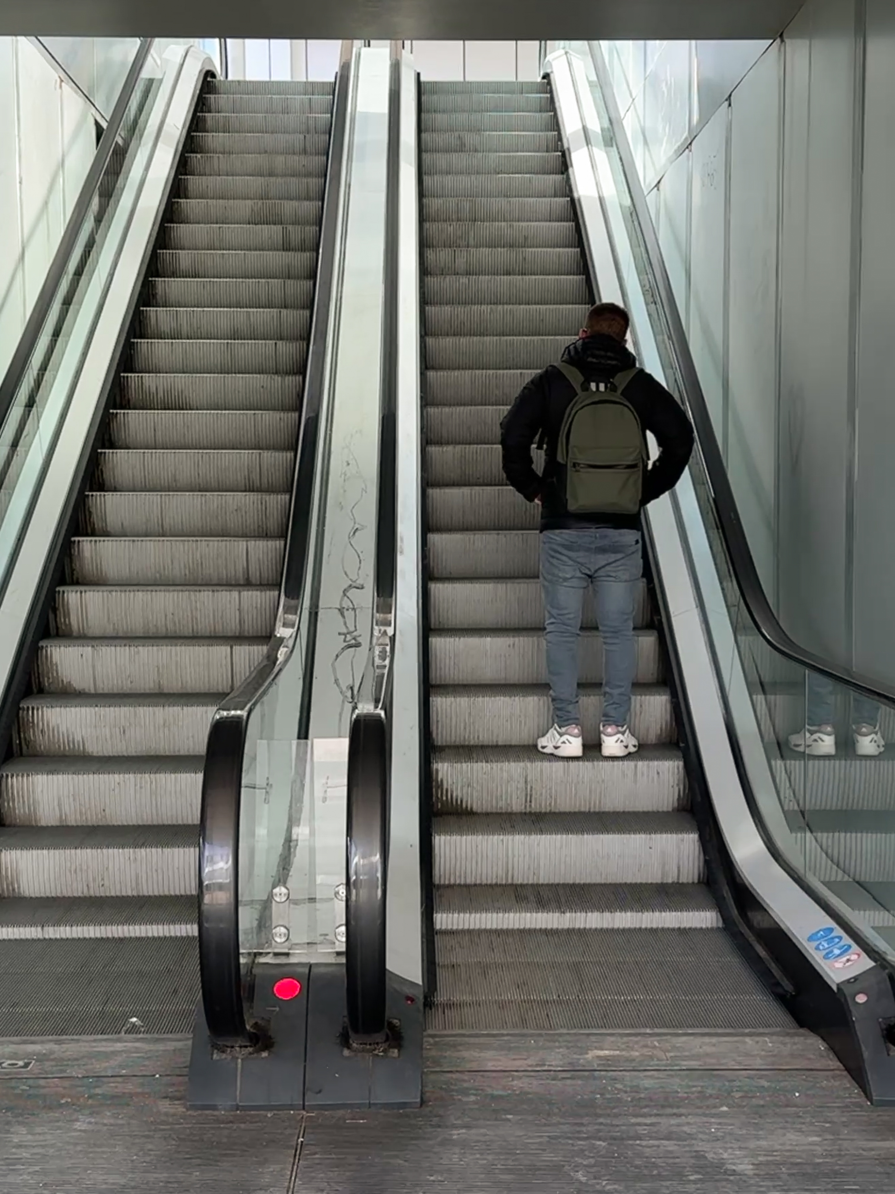 The image shows a man standing on a escalator in an urban setting. He wears a black jacket, jeans, white sneakers, and a green backpack. To his left the is another escalator. The scene has a modern, industrial aesthetic with glass and metal railings, and the escalator appears slightly worn, with visible dirt on the steps.