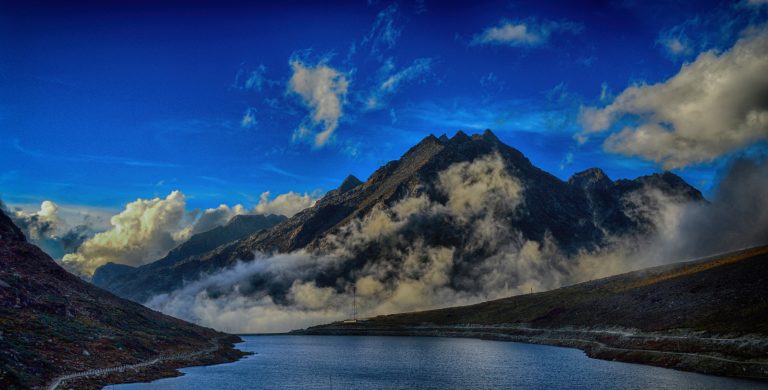 A stunning landscape featuring a serene mountain lake surrounded by rugged peaks, partially covered in mist. The deep blue sky is dotted with wispy clouds, adding depth to the dramatic scenery. A winding road follows the lake’s edge, leading towards the towering mountains in the background.