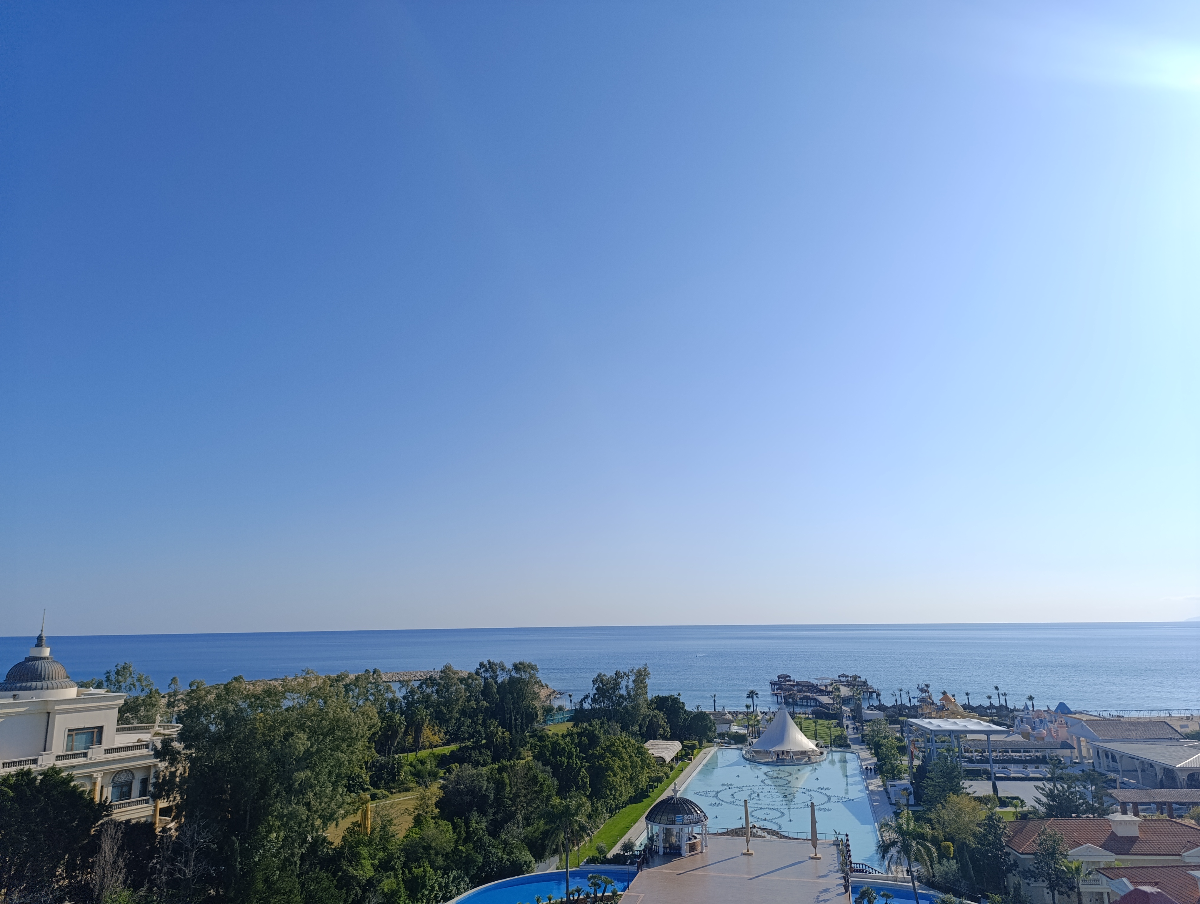 Coastal view with large pool, trees, buildings; clear sky and expansive sea to horizon.