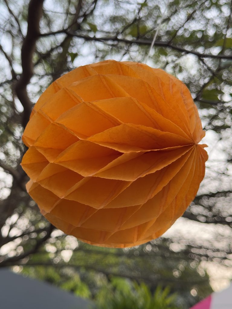 A bright orange paper ball hanging from a tree, serving as a decorative element in an outdoor environment.