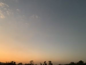 A tranquil scene showing a gradient sky at sunset with shades of orange transitioning to blue, silhouetted trees along the horizon, and a few scattered clouds