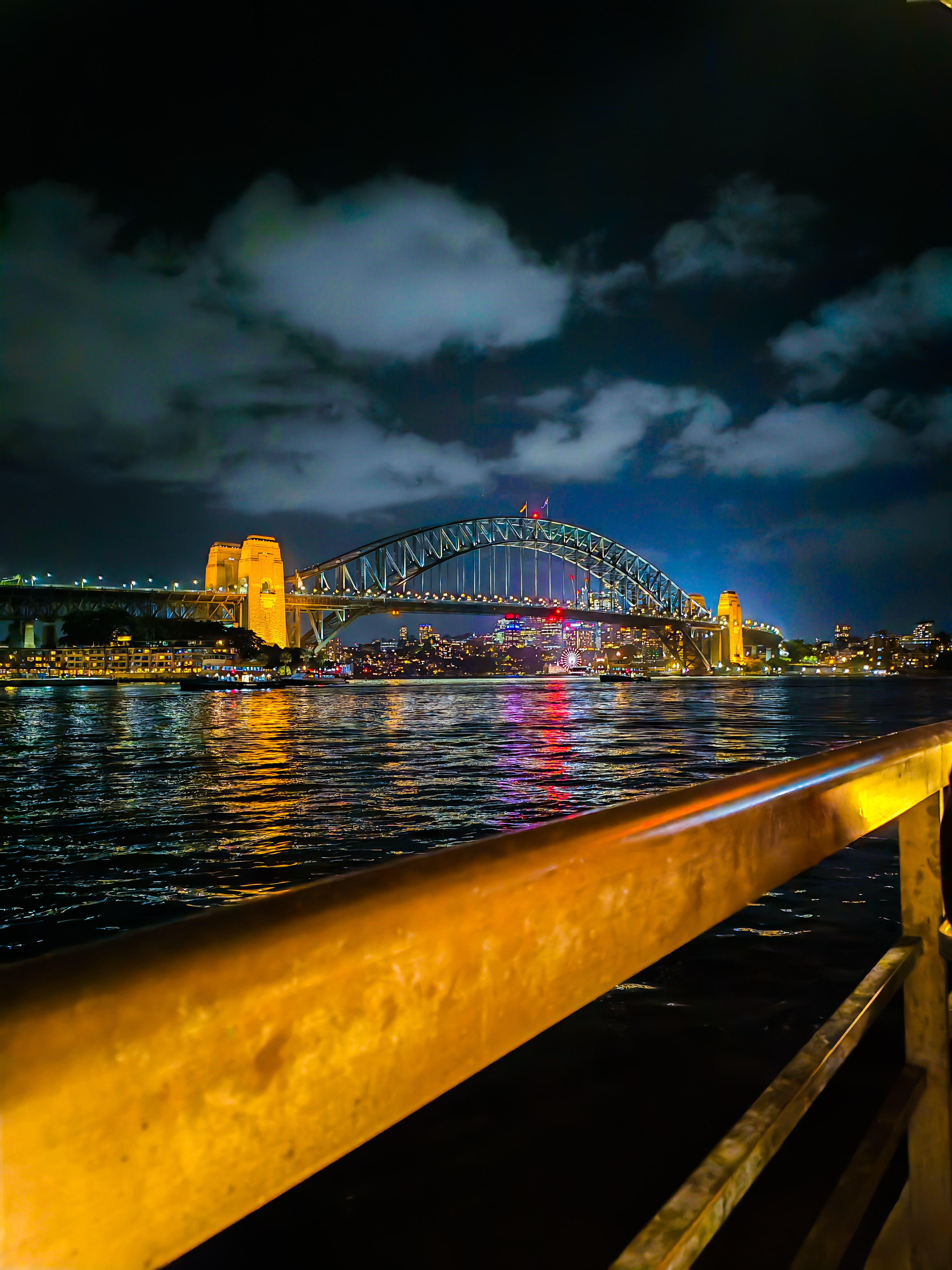 A night view Sydney Harbor Bridge bridge illuminated with lights, reflecting on the water below. The sky is partially cloudy, and there is a wooden railing in the foreground.