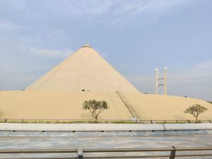 A large, beige, cone-shaped building with a flat base and a small antenna at its peak. 