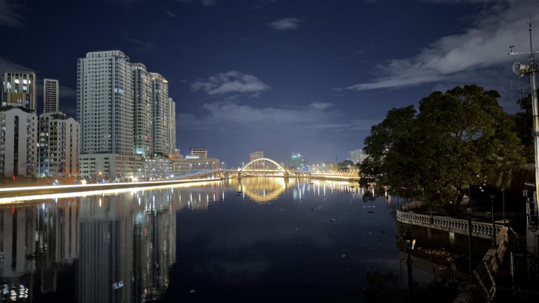 A nighttime cityscape featuring high-rise buildings along a river, with their reflections visible in the water. A well-lit arched bridge spans the river, and trees are visible on one side, adding greenery to the scene.