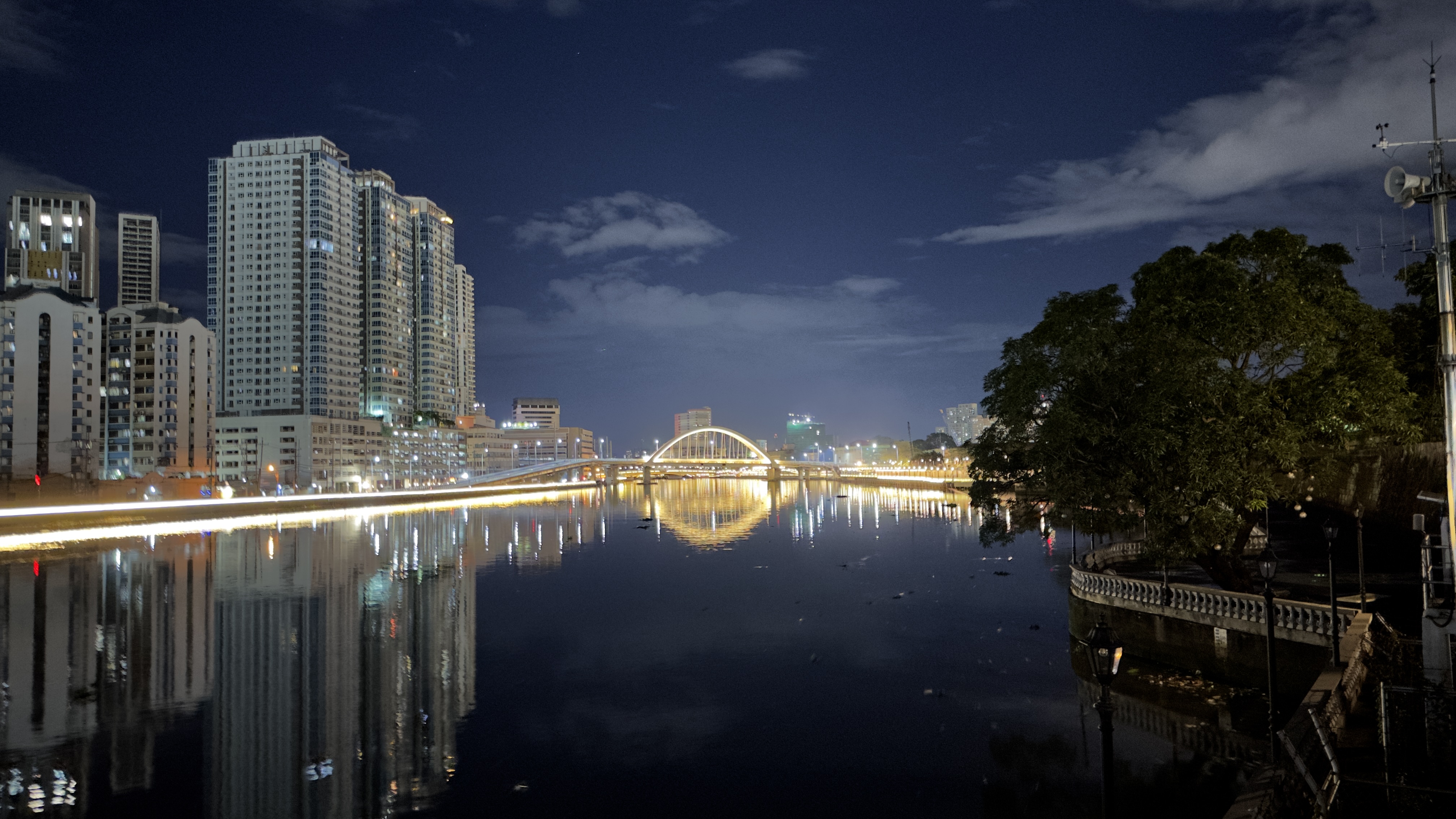 A nighttime cityscape featuring high-rise buildings along a river, with their reflections visible in the water. A well-lit arched bridge spans the river, and trees are visible on one side, adding greenery to the scene. 