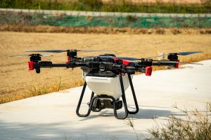 A large drone with four propellers is positioned on a concrete surface, surrounded by dry grass and open fields.
