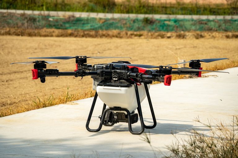 A large drone with four propellers is positioned on a concrete surface, surrounded by dry grass and open fields.