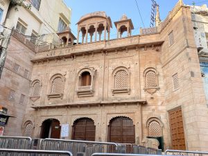 A traditional building with intricate stone carvings and arched windows, featuring a two-story facade with a lattice design and decorative balconies. The structure is made of sandstone and has an ornate, historical architectural style. 