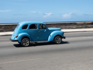 A vintage blue car driving along a coastal road with a stone wall and ocean in the background under a clear blue sky. 