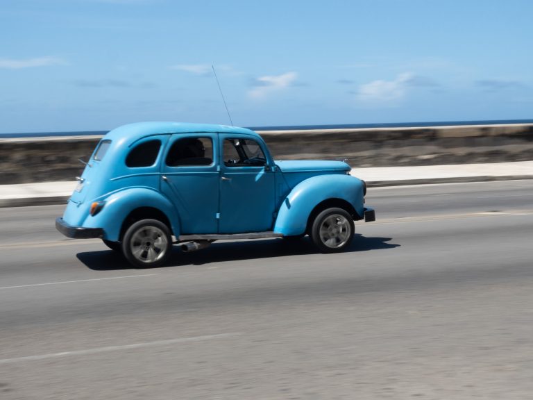 A vintage blue car driving along a coastal road with a stone wall and ocean in the background under a clear blue sky.