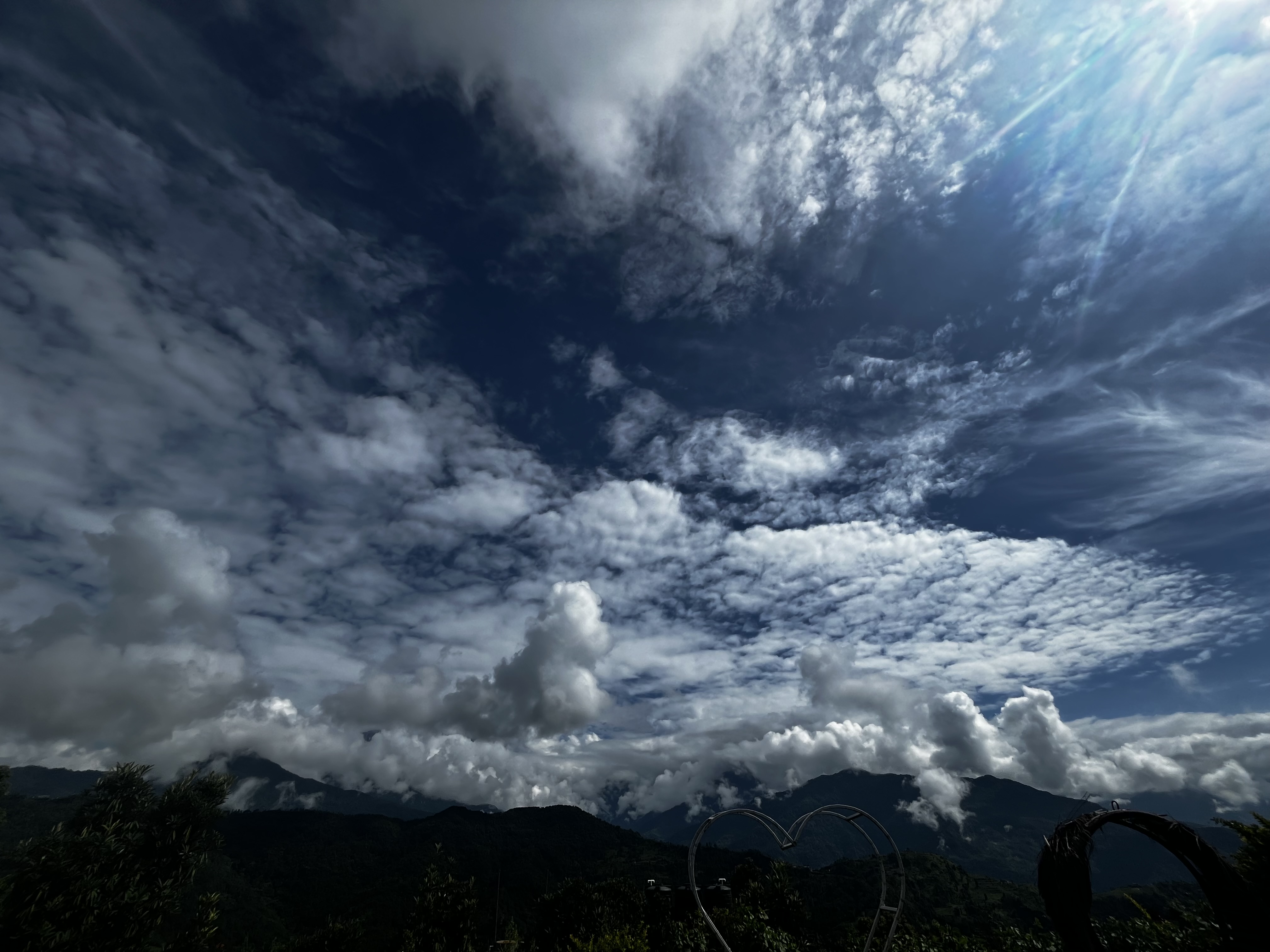 A scenic view of a mountainous landscape under a partly cloudy sky. Fluffy white clouds are scattered across the deep blue sky, with sunlight filtering through. Dark green vegetation and a heart-shaped structure are visible in the foreground.
