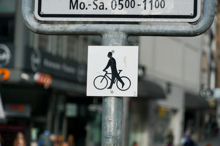 A street sign mounted on a pole, featuring a graphic of a person standing on a bicycle pedal. Above it, there is another sign displaying affected hours. The background shows an urban setting with some blurred figures and shop signage.