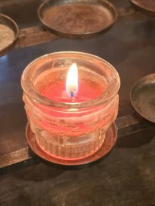 A lit red candle in a clear glass holder, surrounded by other candle holders, some empty. The candle is partially burned, with a visible flame, and is placed on a metal rack or stand.
