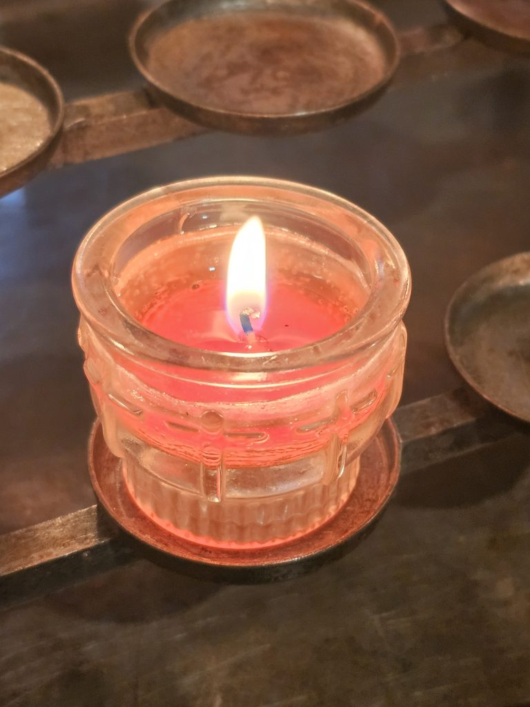 A lit red candle in a clear glass holder, surrounded by other candle holders, some empty. The candle is partially burned, with a visible flame, and is placed on a metal rack or stand.