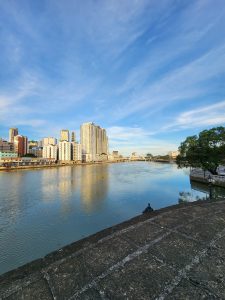 Intramuros, Manila, Philippines - A river with calm water reflecting a clear blue sky and several tall buildings along the opposite bank. A bridge is visible in the distance, and trees line the right side of the image. A paved walkway runs alongside the river in the foreground.