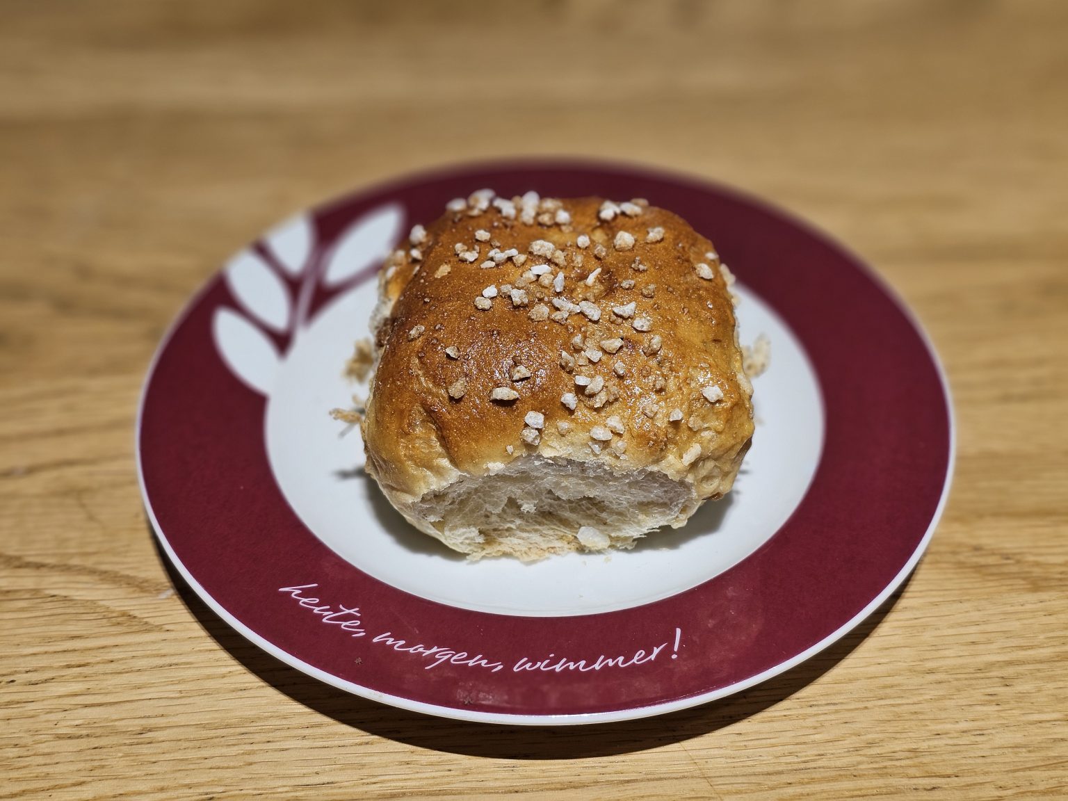 A close-up of a bread roll with a golden brown crust topped with coarse salt, placed on a decorative red and white plate with writing on the rim, on a wooden table.