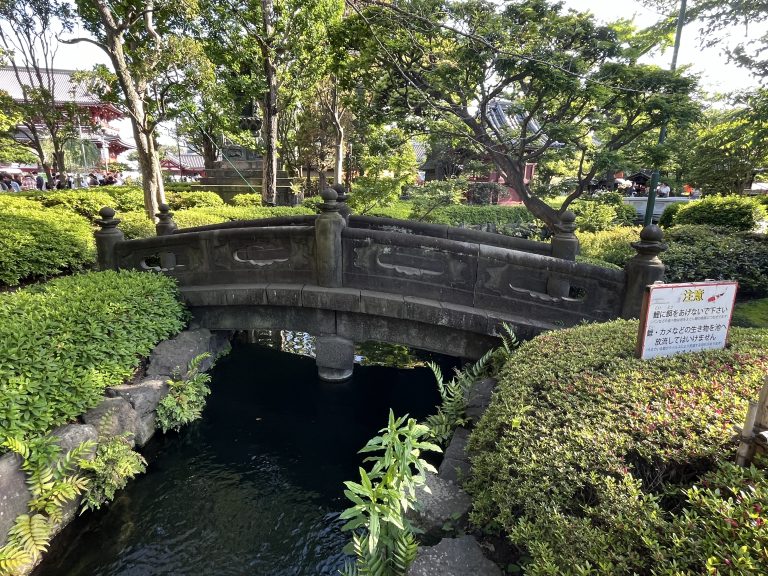 A serene garden scene featuring a traditional stone bridge over a calm stream, surrounded by lush greenery and trees