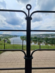 A long view of the Vltava River in Prague, Czech Republic, as seen through a black metal railing of Vyšehrad fort. The river flows through the center of the frame, flanked by green trees and buildings on either side.