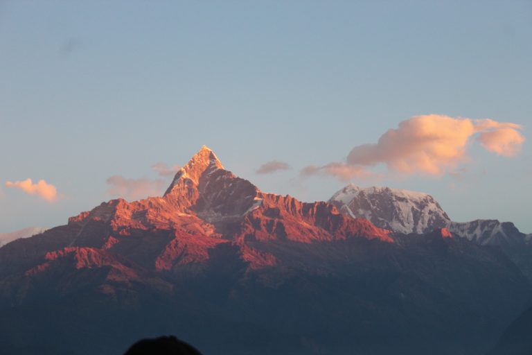 A majestic mountain peak at sunrise with a snow-capped summit, surrounded by rugged terrain. The mountain is bathed in warm sunlight, and a few fluffy clouds float in the blue sky above.