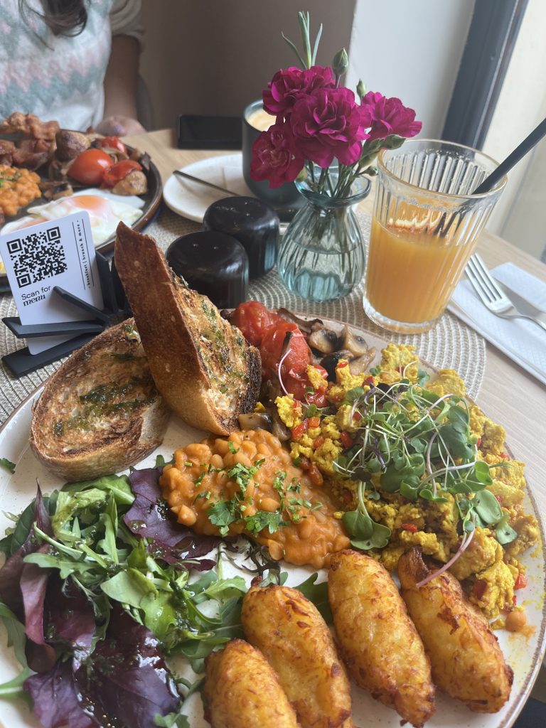 A colorful brunch plate featuring toasted bread with herbs, scrambled tofu with microgreens, baked beans, a fresh green salad, and crispy potato cakes. A glass of orange juice with a straw and a vase with pink flowers are in the background, alongside pepper shakers and a smartphone.