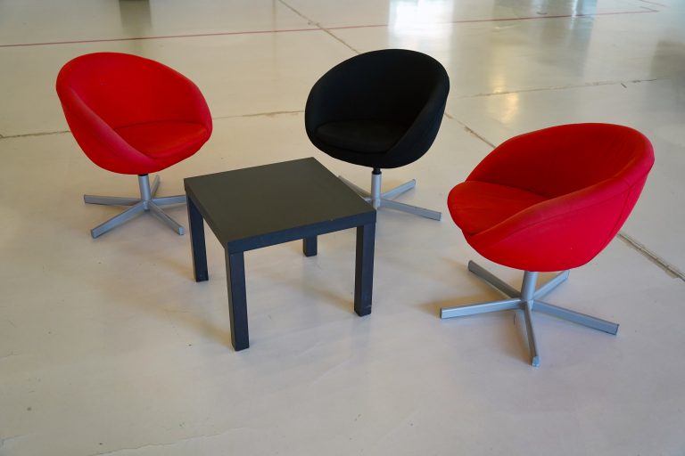 Three modern, round swivel chairs, two red and one black—are arranged around a small, square black table on a light-colored floor.