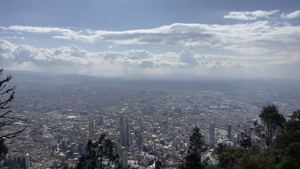 Panoramic view of a large cityscape with numerous buildings under a partly cloudy sky, seen from an elevated vantage point surrounded by trees.