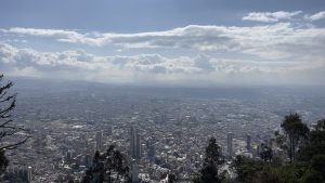 Panoramic view of a large cityscape with numerous buildings under a partly cloudy sky, seen from an elevated vantage point surrounded by trees.