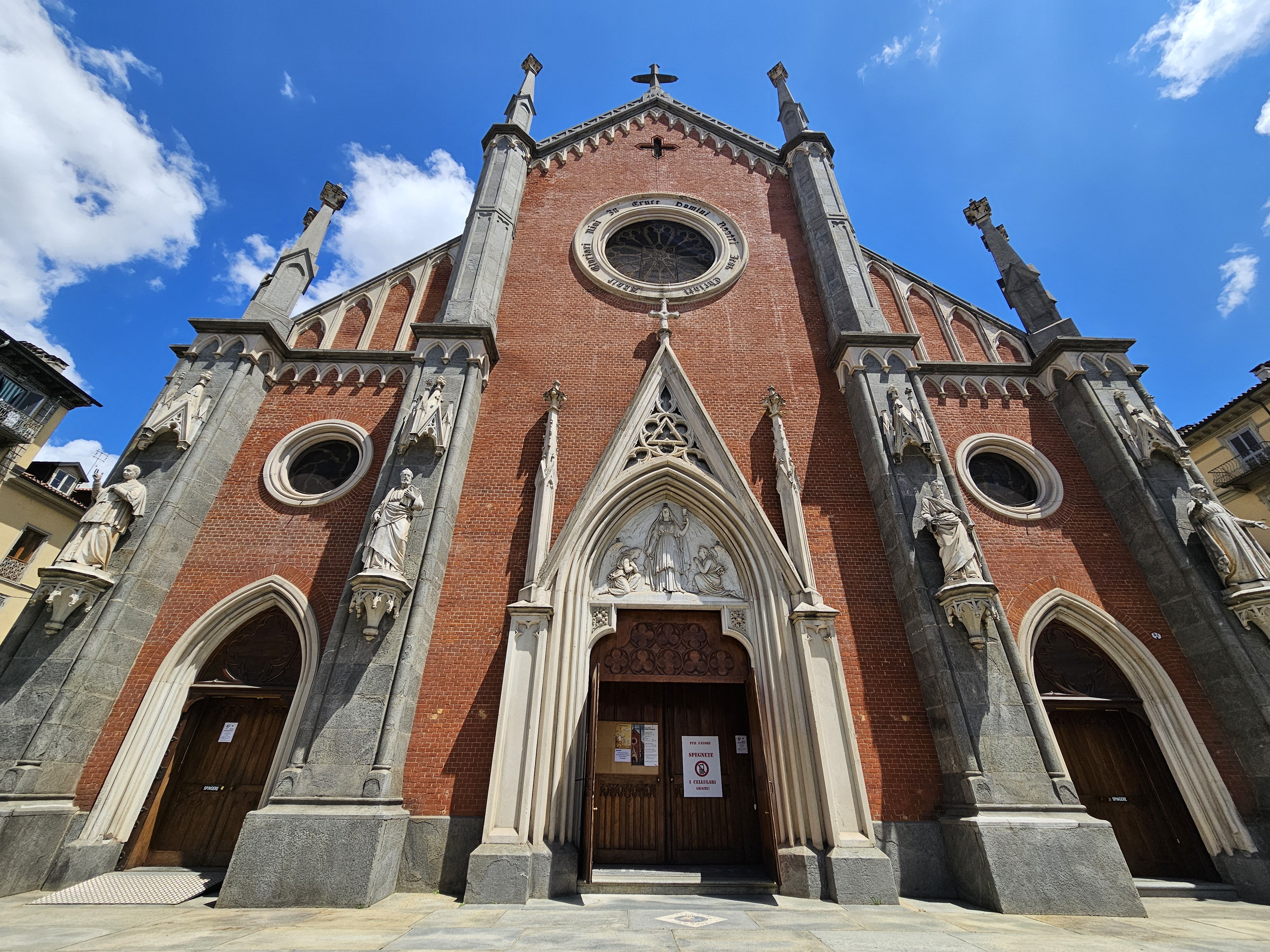 The facade of the Church of Santa Giulia in Turin, Italy, features a red brick exterior with grey stone accents and sculptures. The church's entrance is framed by an ornate archway, and a large circular window is visible above. The architectural style appears to be Gothic Revival, with pointed arches and decorative spires.