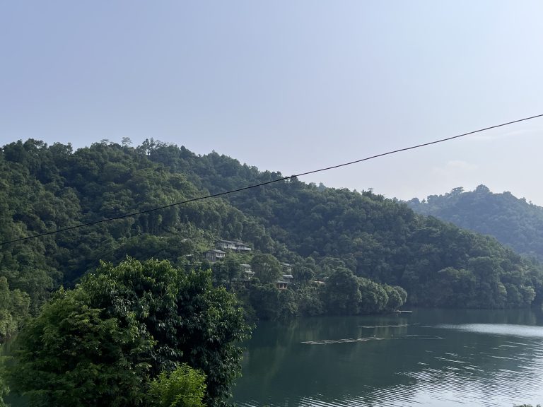 A serene landscape featuring a lush, green hillside with dense forest overlooking a calm body of water. Buildings are nestled in the greenery, and a thin cable runs across the sky. The water reflects the surrounding vegetation under a clear blue sky.