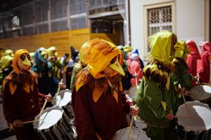  People dressed in colorful costumes resembling birds, with detailed masks and feathered hoods, participating in a parade while playing drums at night during Fasnacht in Basel