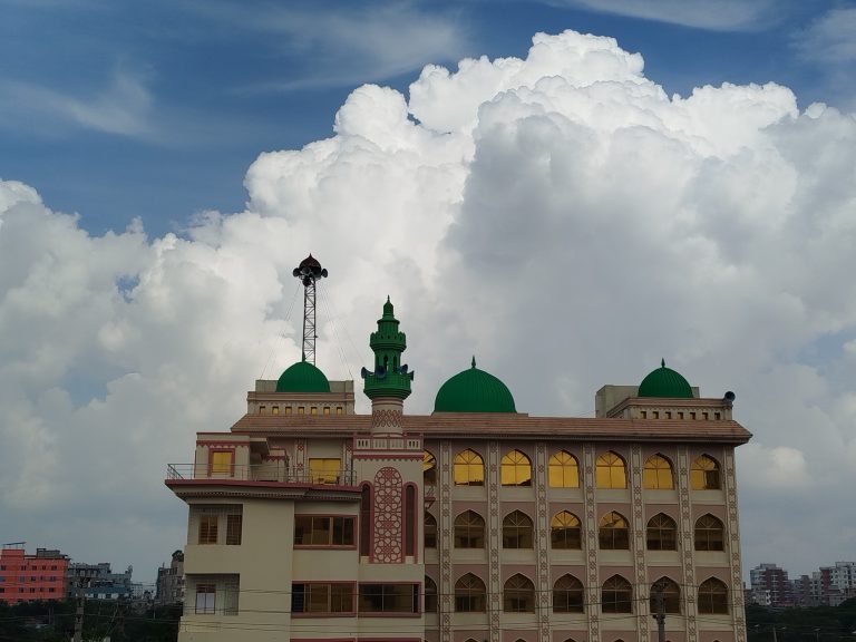 A multi-story mosque building with green domes and golden arched windows, set Against a dramatic backdrop of towering white clouds and blue sky.