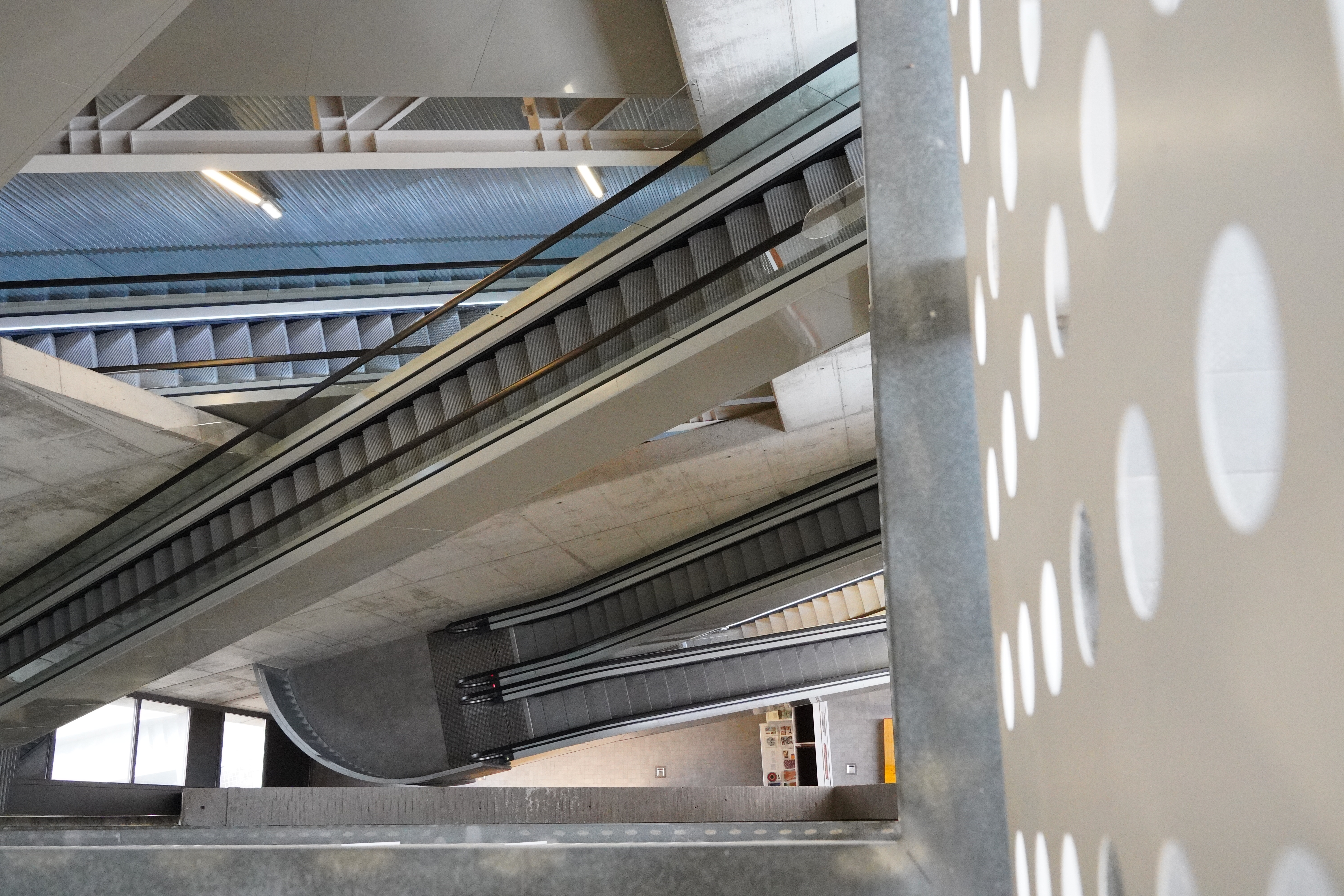 View of multiple escalators inside a modern building, showing intersecting angles and architecture with a perforated wall panel on the right.