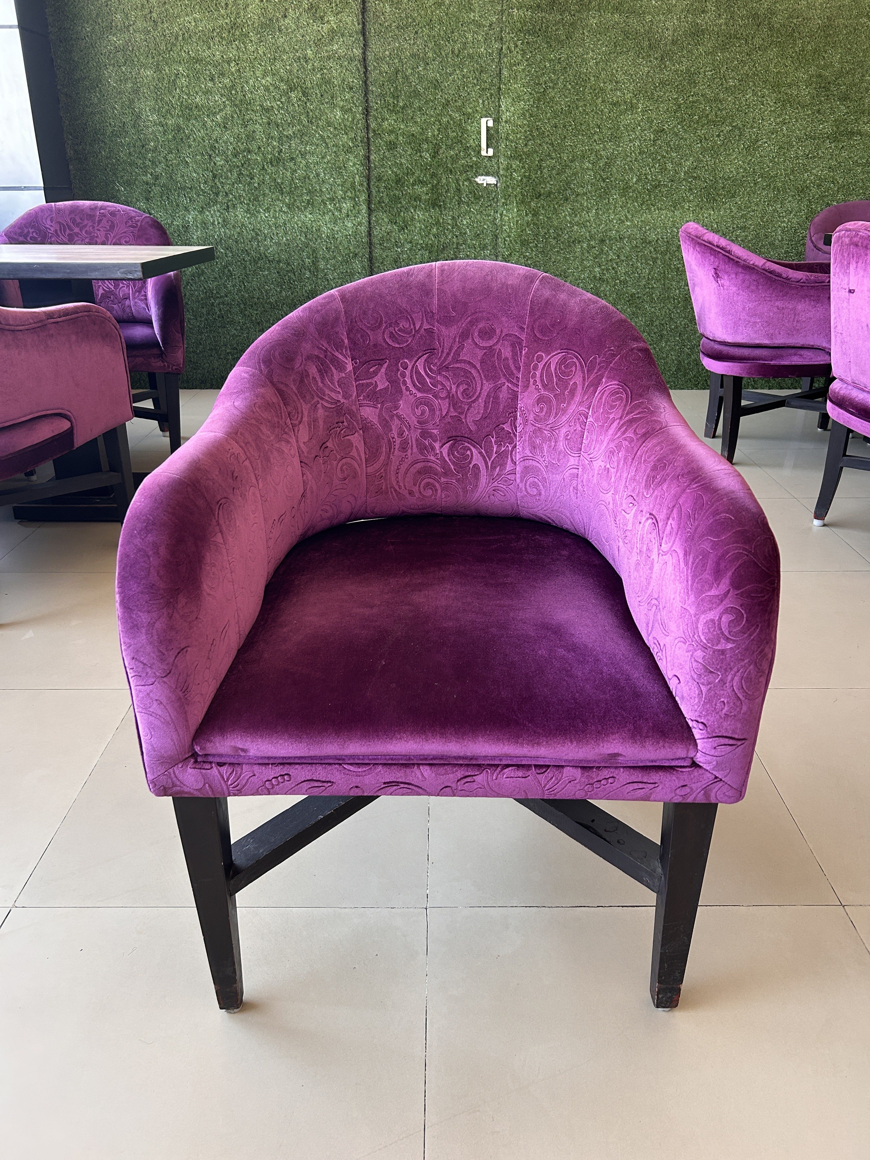 A plush purple armchair with a floral pattern is placed on a tiled floor in an indoor setting. The background features a green textured wall and additional matching chairs around tables.
