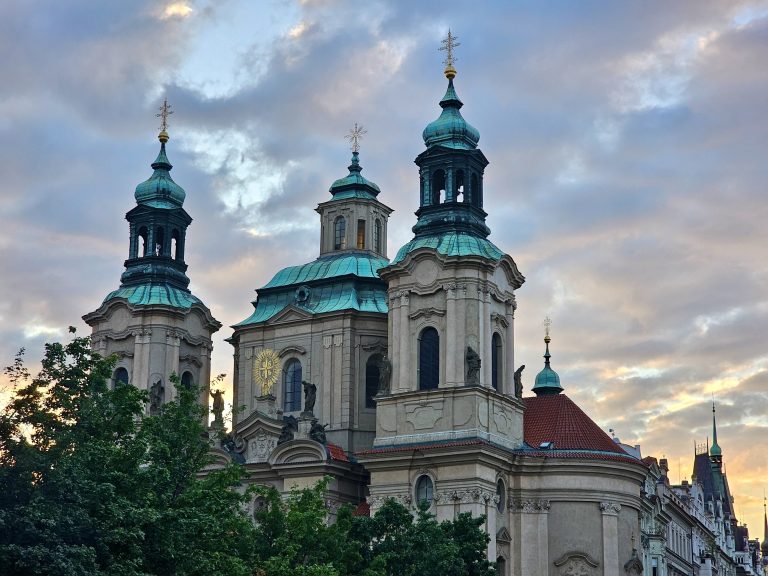 An long view of St. Nicholas Church in Prague (in Old Town Square) in the evening. The Baroque church features three prominent green-domed towers topped with crosses, set against a cloudy sky at dusk.