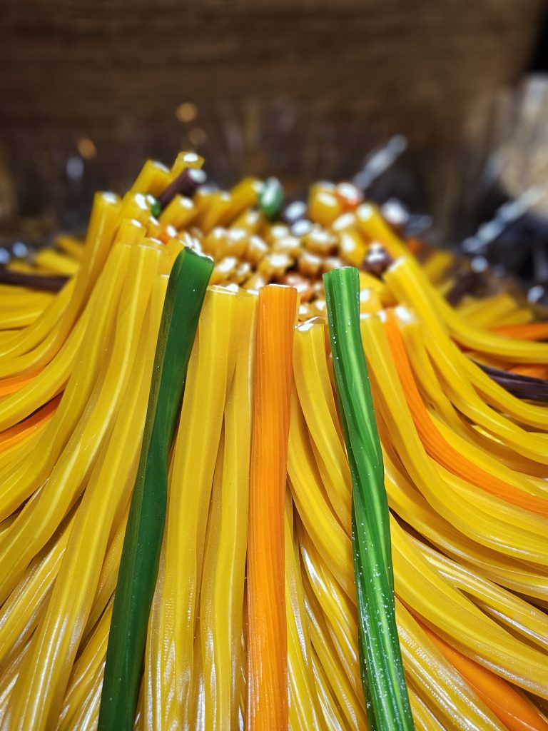 A close-up of a pile of long, colorful rainbow twists gummy candies. The candies are predominantly yellow, with interspersed green and orange pieces, creating a vibrant and textured display.