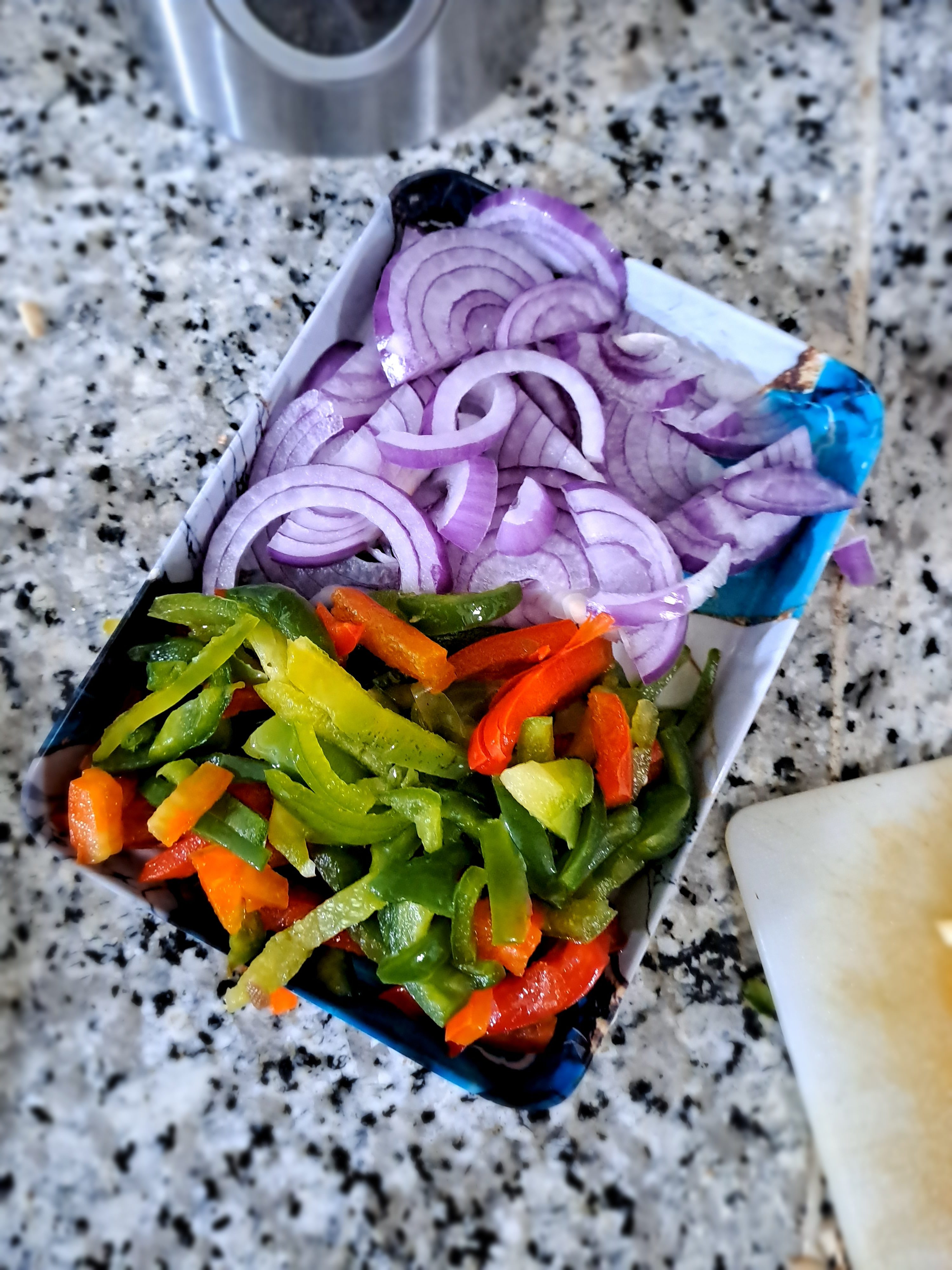 A container on a granite countertop holds sliced red onions and mixed bell peppers.