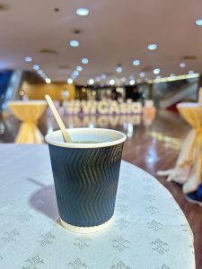 A black disposable coffee cup with a wooden stirrer is on a white patterned tablecloth. In the blurred background,
